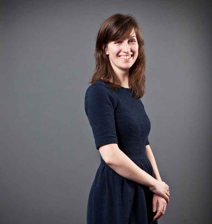 Lecturer with dark brown hair and a grey backdrop smiling at camera